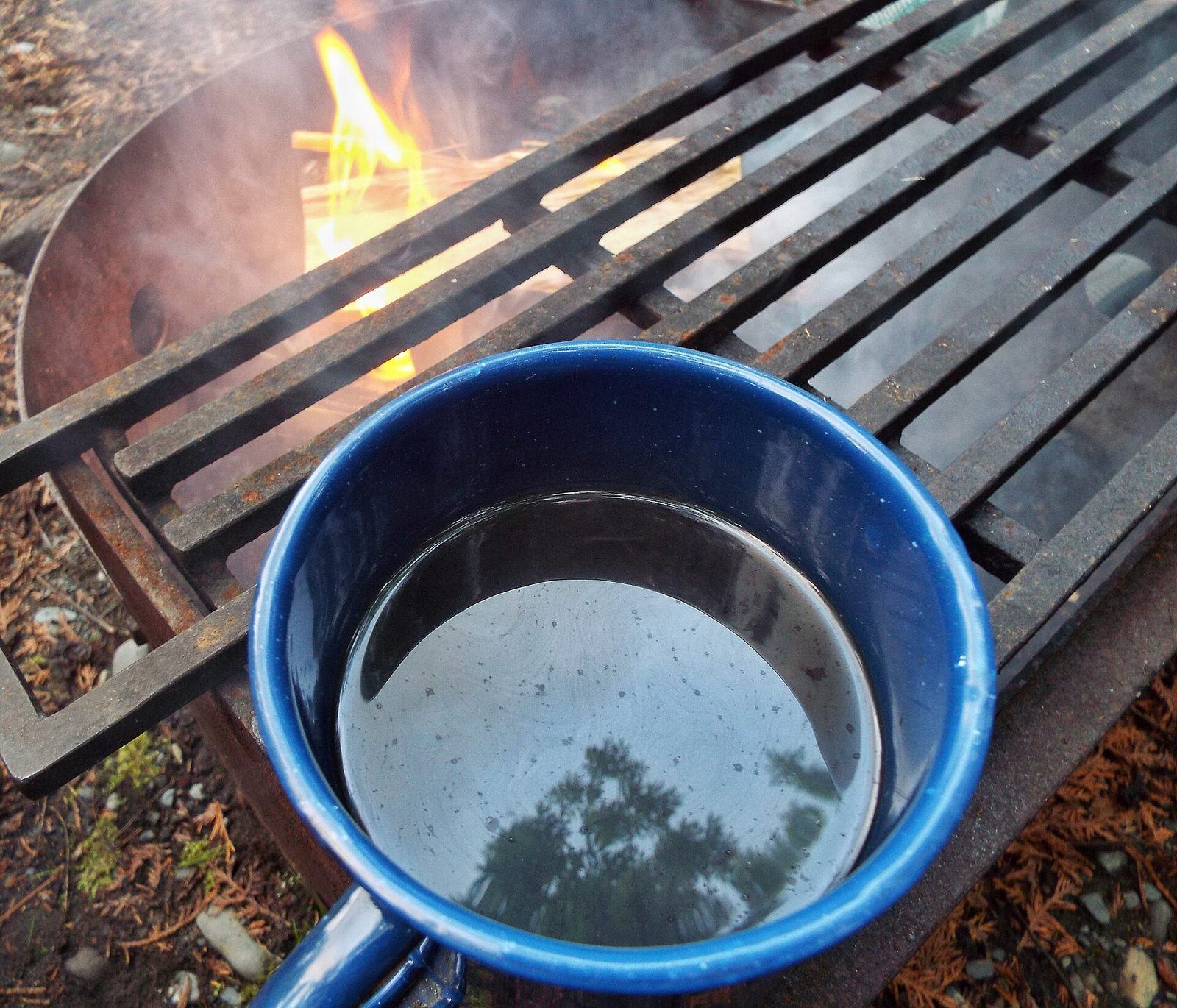 A blue enamel mug sitting on the iron grate of a campfire ring with flames burning behind it