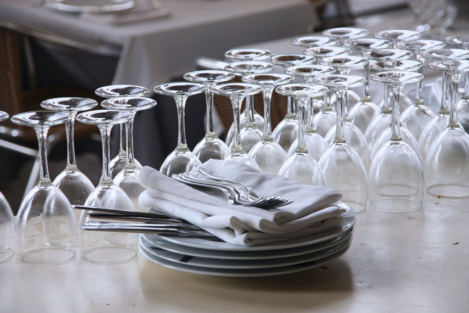 Rows of wine glasses standing upside down on a white restaurant table beside a stack of plates