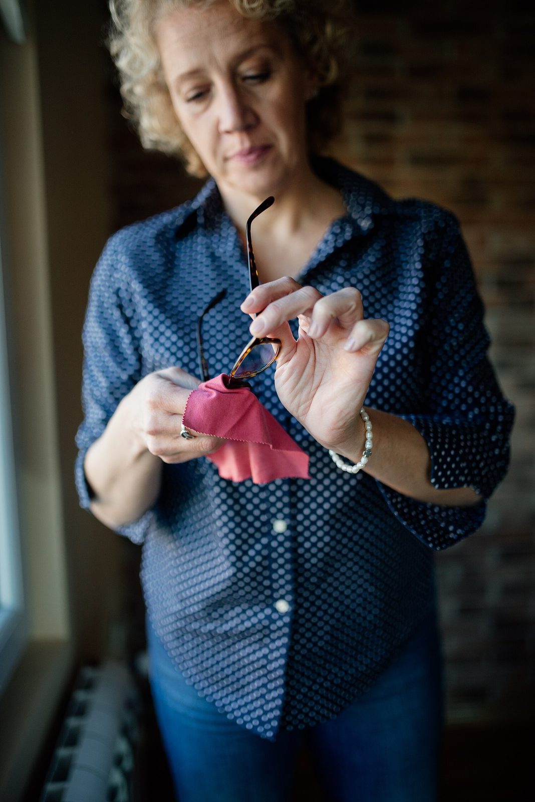 A woman standing by a window holding her eyeglasses up to check the lenses