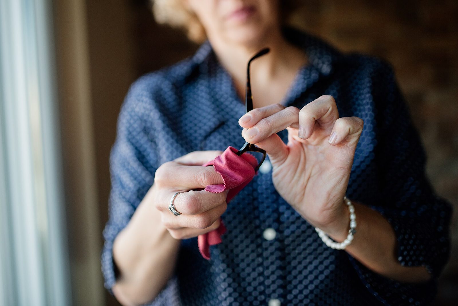 A woman holding her folded eyeglasses in one hand and wiping a lens with a cloth