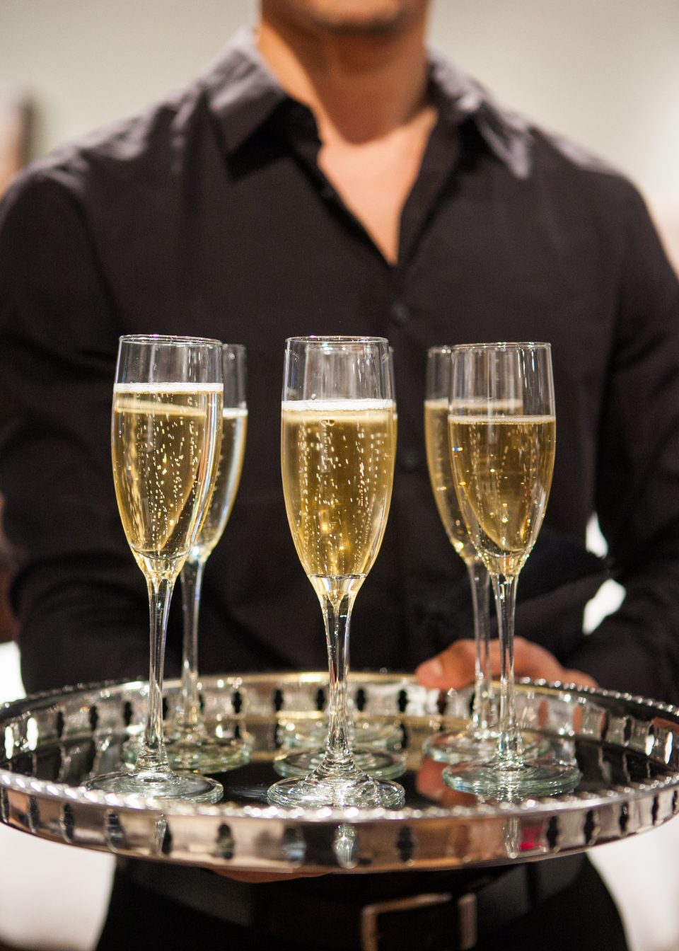 A waiter in a dark shirt holding a round silver tray carrying four champagne glasses